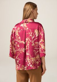 Blusa in satin di un vivace rosa con motivo floreale dorato. Maniche a tre quarti, vestibilità rilassata e dettagli di pieghe sottili sul retro.