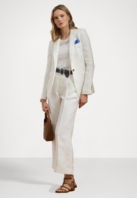 White linen suit with wide-leg pants and a fitted blazer. Sheer, textured top underneath. Brown leather handbag and strappy sandals. Blue pocket square.