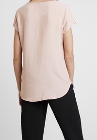 Jasnoróżowa bluzka z krótkim rękawem, okrągłym dekoltem, gładką fakturą i lekko zaokrąglonym dołem. W zestawie z czarnymi spodniami.