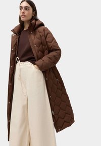 Brun kviltet pufferjakke med hætte; har trykknaplukning og sidelommer. Båret over en mørkebrun skjorte og lysecreme bukser med vidde.