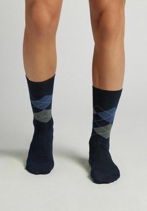 Des jambes portant des chaussettes bleu marine avec un motif argyle bleu et gris, se tenant sur une surface gris clair.