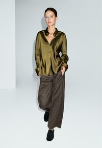 Camicia di raso verde oliva con colletto e bottoni frontali, abbinata a pantaloni marroni larghi. Il modello indossa mocassini neri e ha le mani nelle tasche.