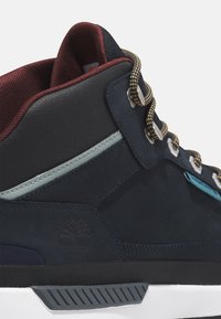 Zapatilla azul marino con una parte superior de cuero y ante, cuello acolchado, acentos en contraste y ganchos de metal para los cordones. Presenta una suela de goma con agarre.