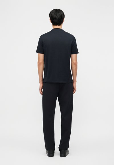 Άνδρας που στέκεται πλάτη, φοράει απλό μαύρο κοντομάνικο πουκάμισο, μαύρο παντελόνι και μαύρα παπούτσια μπροστά σε λευκό φόντο.