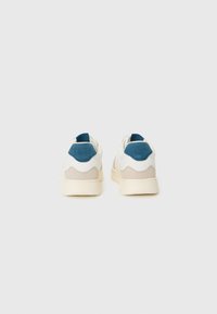 Un par de zapatillas blancas con detalles en beige y azul marino, vistas desde la parte trasera sobre un fondo blanco liso.