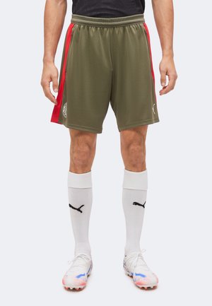 Homme portant un short de sport vert olive et rouge, des chaussettes blanches Puma et des crampons blancs avec des touches bleues et orange, debout devant un fond uni.