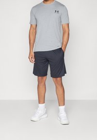 Hombre vistiendo una camiseta gris de Under Armour, pantalones cortos deportivos negros, calcetines blancos y zapatillas blancas de pie con una mano en el bolsillo.
