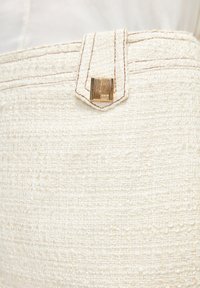 Gros plan d'un tissu à texture crème avec passant de ceinture cousu et petit bouton carré en métal doré.