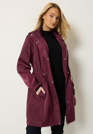 Femme blonde portant une longue veste bordeaux à capuche sur une tenue noire, posant avec une main dans la poche sur un fond uni.