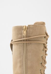 Botín de tobillo de ante beige con una cremallera visible y cordones en el lateral. Presenta ojales y una superficie suave y texturizada.