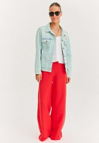 Jasnoniebieska kurtka dżinsowa, biała crop top i szerokie czerwone spodnie. Okulary przeciwsłoneczne i naszyjnik dopełniają stylizację, charakteryzującą się swobodnymi, żywymi kolorami.