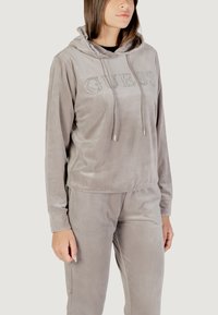 Sudadera de felpa gris con cordón, que presenta un logo decorativo de "GUESS" en rhinestones. Combinada con pantalones ligeros a juego.