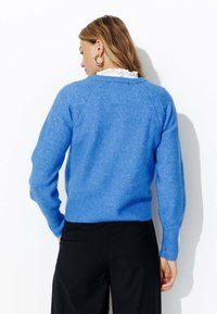 Pull en tricot bleu avec un col rond, des poignets et un ourlet côtelés, présentant un design texturé sur les épaules. Porté par-dessus un pantalon sombre.