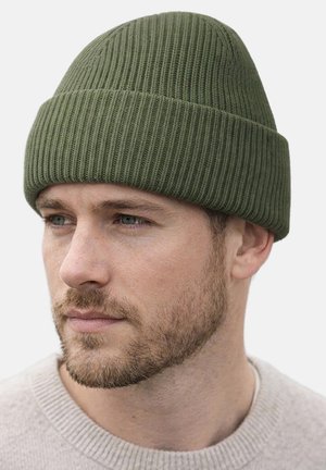 Man with short beard wearing olive green ribbed knit beanie and light gray sweater, looking to the left on plain light background.