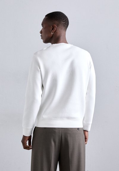 Sweatshirt de algodão branco com punhos canelados, decote redondo e textura suave, combinado com calças cinzas. Foco na vista traseira e design minimalista.