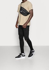Svarta sneakers, svarta trasiga jeans, beige t-shirt och en svart axelremsväska. Outfiten har en avslappnad streetwear-stil.