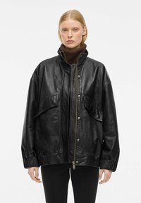 Veste en cuir noir avec un col montant, une fermeture éclair et deux poches avant. Caractérisée par une coupe décontractée et des poignets froncés pour une texture douce.