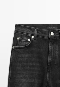 Jean en denim noir affichant la partie supérieure avant avec la ceinture, un bouton en métal, des passants de ceinture et deux poches avant avec rivets.