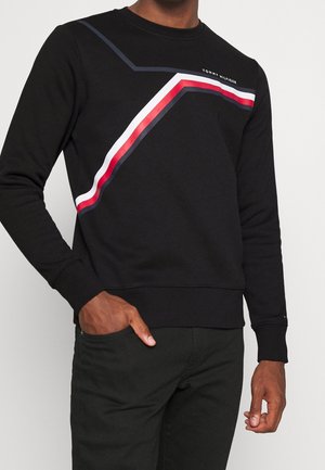 Muž v černé mikině s diagonálními červenými, bílými a námořnickými pruhy a nápisem "Tommy Hilfiger", stojící s rukou v kapse na jednoduchém pozadí.