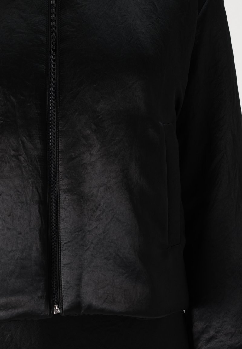 Veste noire courte en matériau texturé avec fermeture éclair sur le devant et poche latérale. Fini doux, léger éclat et design minimaliste.