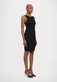 Black, sleeveless sheath dress with a fitted silhouette, smooth texture, and round neckline. Worn with black stiletto heels.