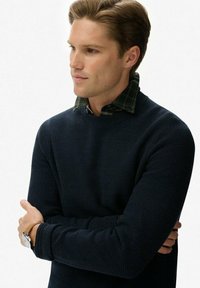 Marineblauer Strickpullover, der über einem grün karierten Hemd getragen wird. Der Pullover hat einen Rundhalsausschnitt und ein strukturiertes Material. Als Zubehör wird eine Uhr getragen.