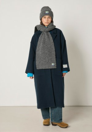 Jūrinis vilnonis paltas su didelėmis kišenėmis, pilka raukšlėta šalikėlis, mėlynas megztinis, pilkas skrybėlė ir rudi mediniai šlepetės, prieš neutralią foną.