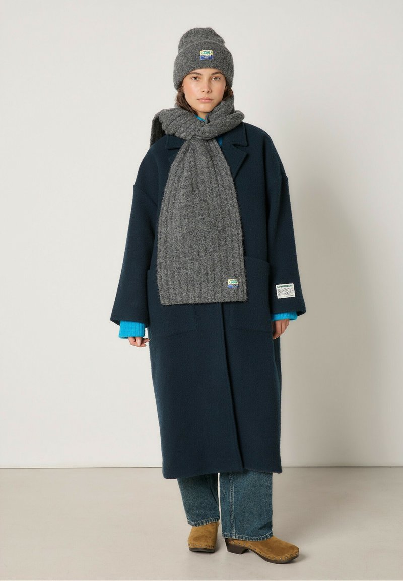 Marineblå uldfrakke med store lommer, grå ribbet tørklæde, blå sweater, grå hat og brune klipklapper, sat mod en neutral baggrund.