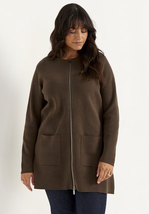 Frau trägt eine braune Strickjacke mit Reißverschluss und vorderen Taschen sowie dunkle Hose, steht vor einem einfarbigen hellen Hintergrund.