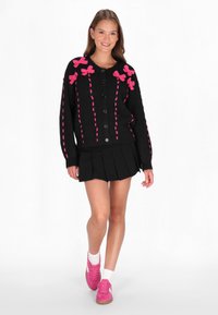 Juodas kardiganas su rožiniais kaspinais, mygtukais priekyje ir rožinėmis siūlėmis. Dėvimas virš sulenktos juodos sijono ir rožinių batų.