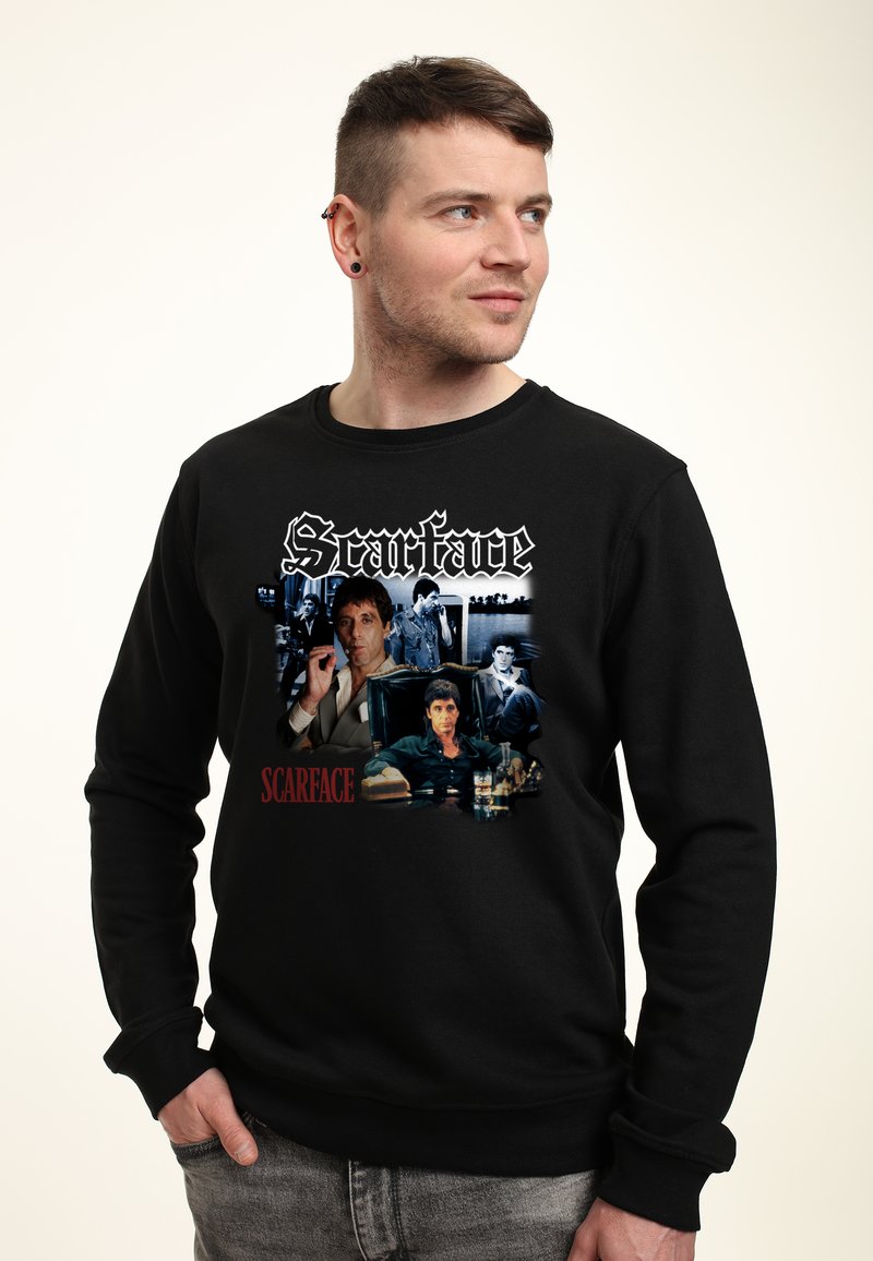 Felpa nera con grafica "Scarface" che presenta immagini dei personaggi e il titolo in bold bianco e rosso. Tessuto morbido e strutturato, vestibilità casual.