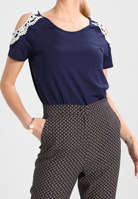 Top azul marino con diseño de hombros al aire y borde de encaje floral; combinado con pantalones de talle alto con estampado floral en una paleta de colores contrastantes.