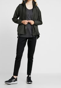 Vandtæt sort jakke med hætte over en mørkegrå skjorte, kombineret med sorte bukser med hvide sidestreger og sorte sportssko.