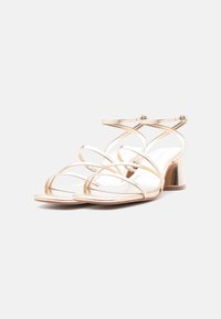 Ζευγάρι μεταλλικά χρυσά πέδιλα με ανοιχτό δάχτυλο, φαρδύ τακούνι και λεπτά διασταυρωμένα λουριά με αγκράφες στον αστράγαλο σε λευκό φόντο.