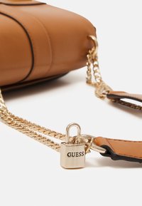 Tan synthetisch lederen handtas met gouden hardware, kettingriem en een hangslotdetail met het label "GUESS." Beschikt over strakke lijnen en contrasterende accenten.