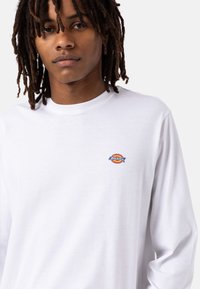 Witte lange mouwen t-shirt van katoen, met een klein multicolored logo op de linkerborst. Eenvoudig, klassiek ontwerp met een ronde hals.