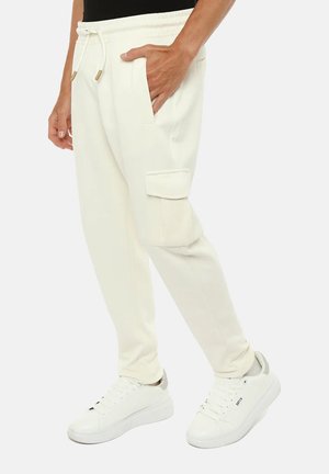 Uomo che indossa pantaloni cargo jogger color avorio con tasca laterale e sneakers bianche basse, in piedi con una mano in tasca.