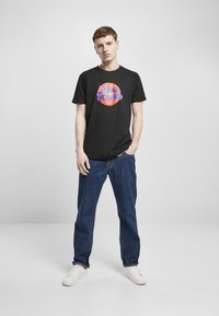 Svart bomull T-skjorte med en fargerik "TUNE SQUAD" logo på en sirkulær grafikk. Brukt med blå denim-jeans og hvite joggesko.