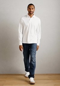 Mann, der drinnen auf einem Holzfußboden geht, trägt ein weißes Langarm-Polohemd, dunkelblaue Jeans und weiße Sneakers.