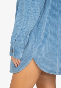 Abito camicia in corda azzurro chiaro con strisce verticali, maniche lunghe, polsini con bottoni, fessure laterali e dettagli a scallop sull'orlo.
