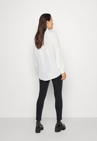 Blusa branca de manga longa com punhos franzidos, combinada com leggings pretas. A blusa tem uma bainha arredondada e uma textura leve e suave.