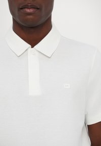 Hvit polo skjorte med strukturert stoff, klassisk krage, to-knappers plaket og en liten, preget logo på brystet.
