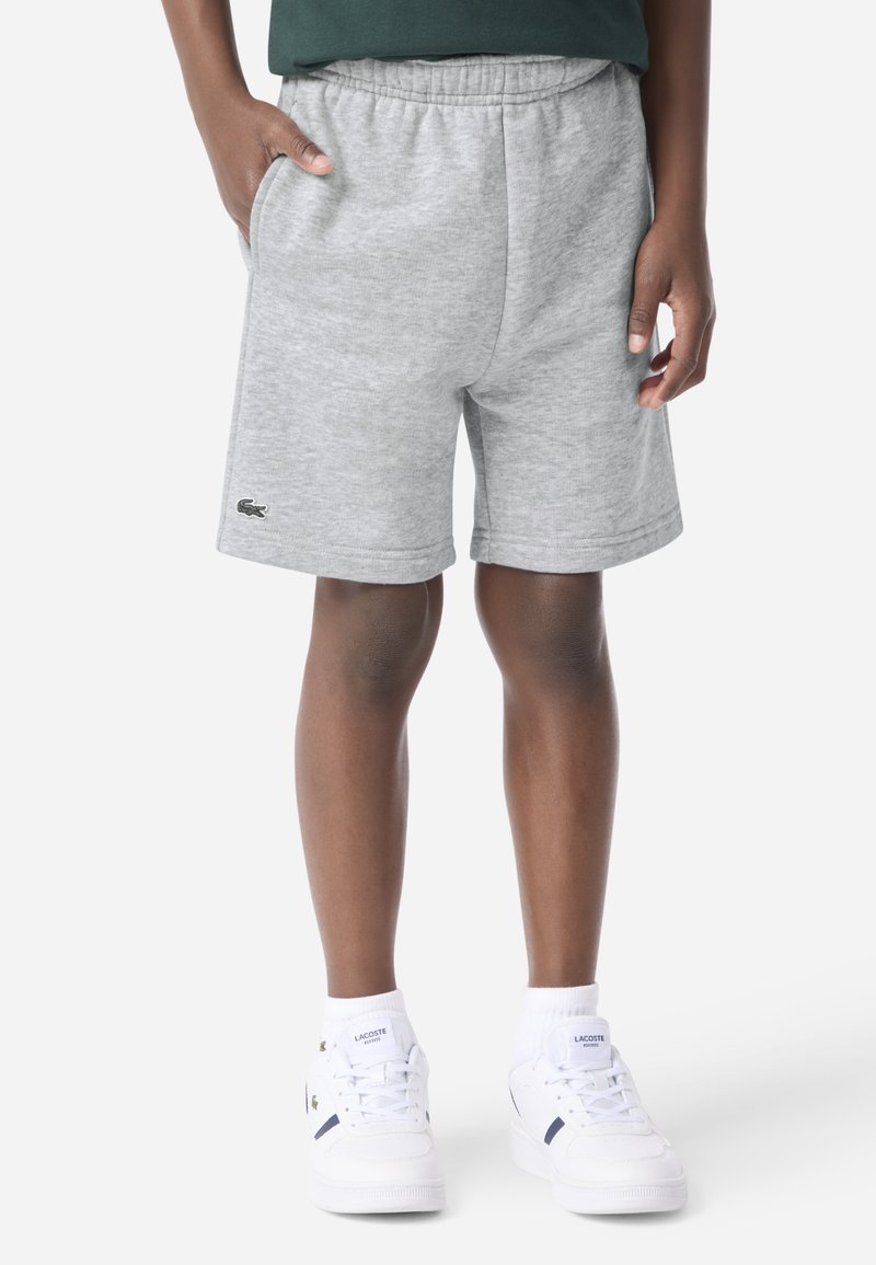 Grå bomuldsblandingsshorts med elastisk talje, sidelommer og Lacoste-logo broderet nær kanten. Kombineret med hvide sneakers.