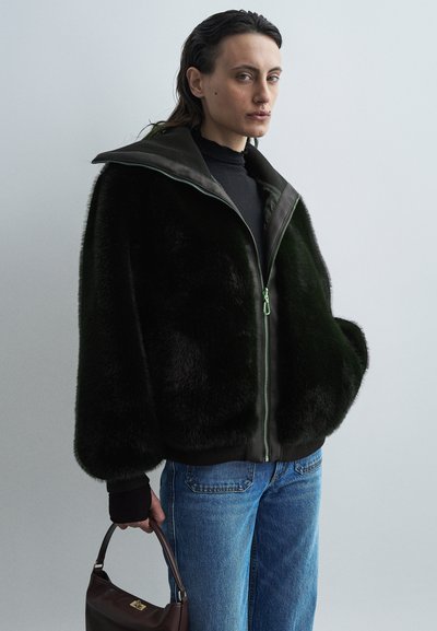 Giacca in pelliccia sintetica di colore verde scuro con colletto alto, chiusura con zip e polsini a coste. Indossata sopra una maglia nera, abbinata a jeans azzurri chiari.