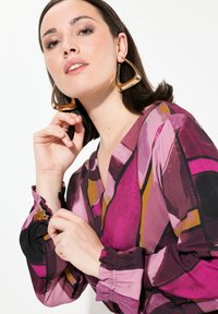 Bunt gemusterte Bluse mit V-Ausschnitt und Puffärmeln, die abstrakte Formen in Pink, Lila und Gelb zeigt. Große geometrische Ohrringe aus Gold.