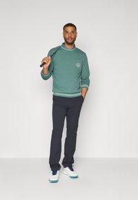 Felpa verde con un piccolo logo sul petto, polsini e orlo a coste. Indossata con pantaloni scuri e scarpe da golf bianche con dettagli blu.