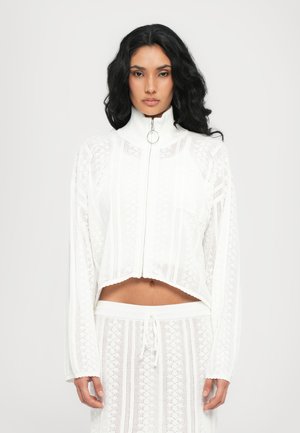 Veste blanche zippée courte avec col haut, texture en tricot ouvert complexe et ourlet discret en forme de vagues, assortie à un pantalon à cordon de serrage.