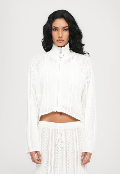 Giacca bianca a zip accorciata con alto colletto, intricata texture a maglia aperta e un sottile orlo scalloped, abbinata a pantaloni con coulisse coordinati.