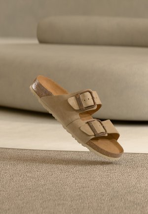Beige Veloursledersandale mit zwei verstellbaren Schnallen, Korkfußbett und strukturierter Gummisohle. Verfügt über einen leicht erhöhten, glänzenden Akzent an der Ferse.