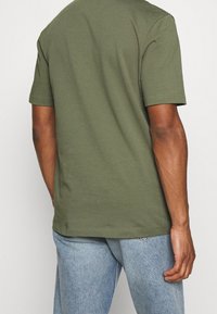 Camiseta de algodón verde olivo con mangas cortas y escote redondo; textura suave y corte relajado, la vista trasera muestra detalles de costura.